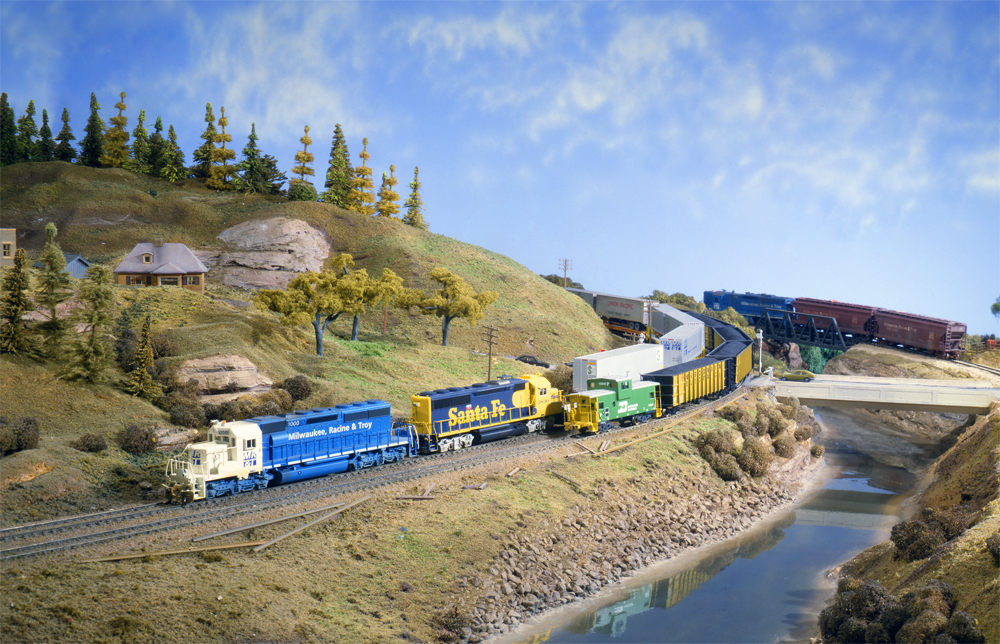 Photo of blue-and-white locomotive on a scenicked model railroad with hills and river.