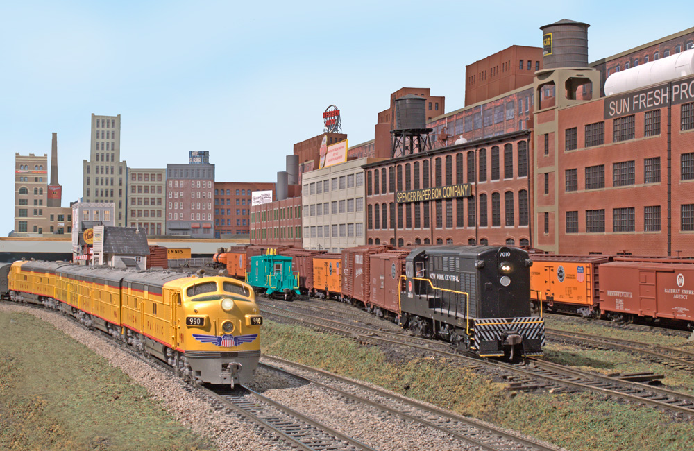 Four yellow diesels curve in front of a black switch engine in front of an industrial urban scene