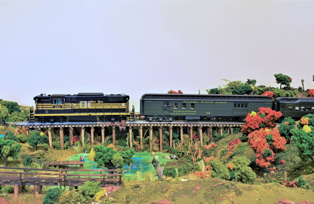 A diesel-powered passenger train rolls over a low wooden trestle behind a colorful park scene