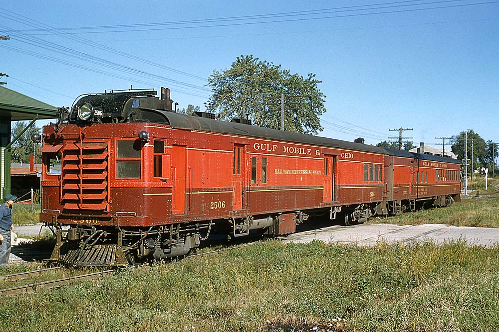 Red and maroon gas motorcar with trailer in front of station