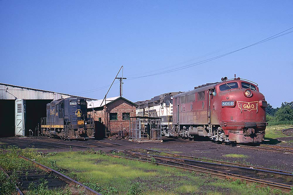 Red streamlined diesel locomotive with two black and white and one blue and yellow diesel locomotives in front of enginehouse
