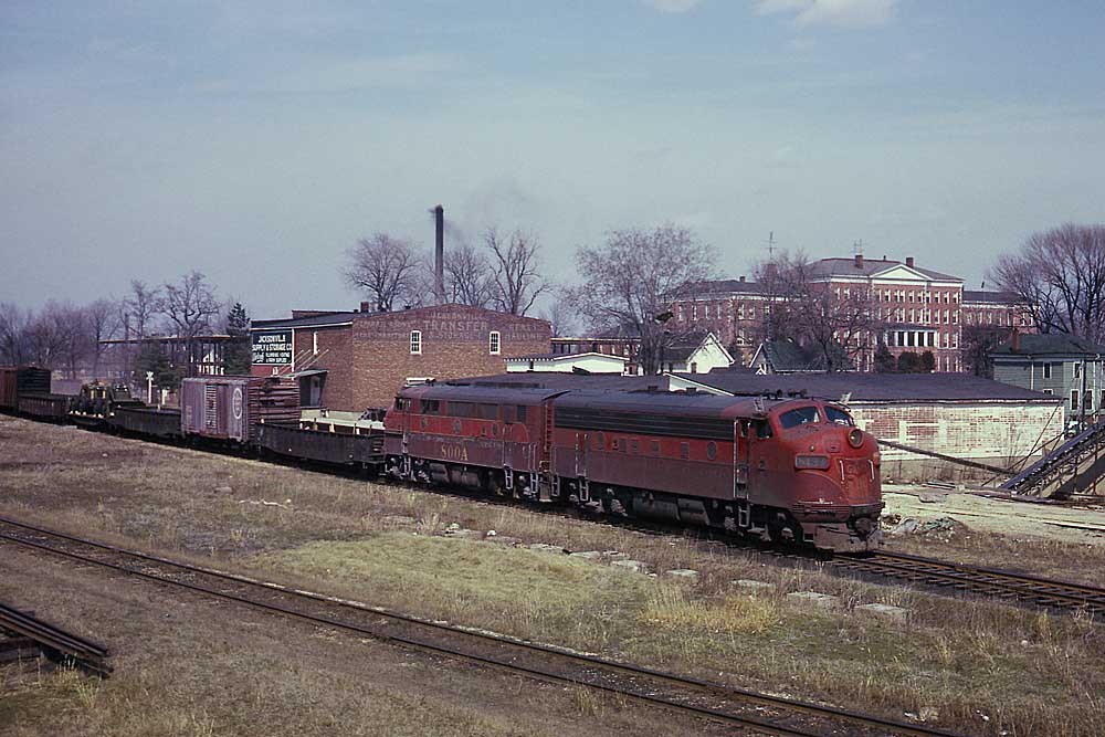 Maroon and red diesel locomotive with freight train in front of brick buildings