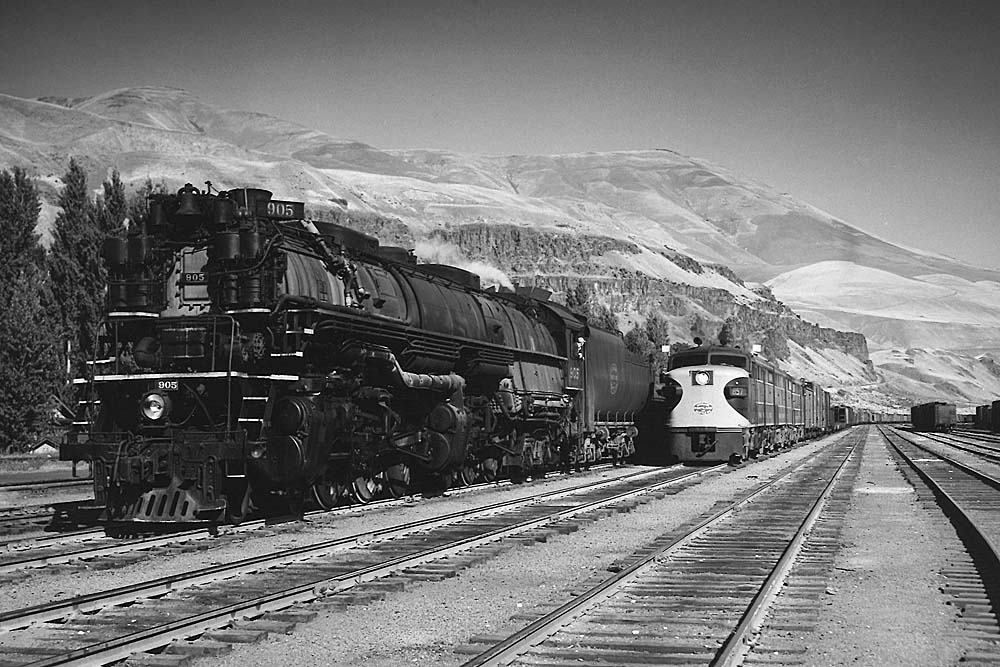 Diesel-powered freight train passes steam locomotive below mountains