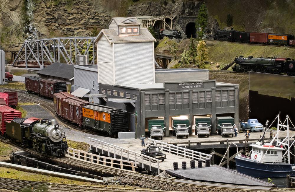 A steam locomotive pulls past a gray waterfront warehouse with a fishing boat docked nearby
