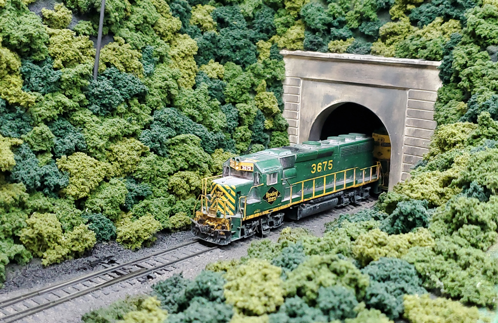 A green-and-yellow hood unit diesel emerges from a tunnel set in a wooded mountain