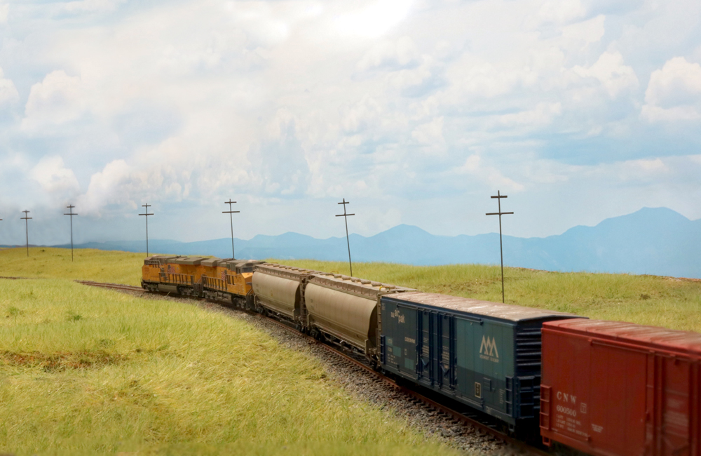 A pair of yellow-and-gray diesels pulls a collection of freight cars through the gently rolling prairie