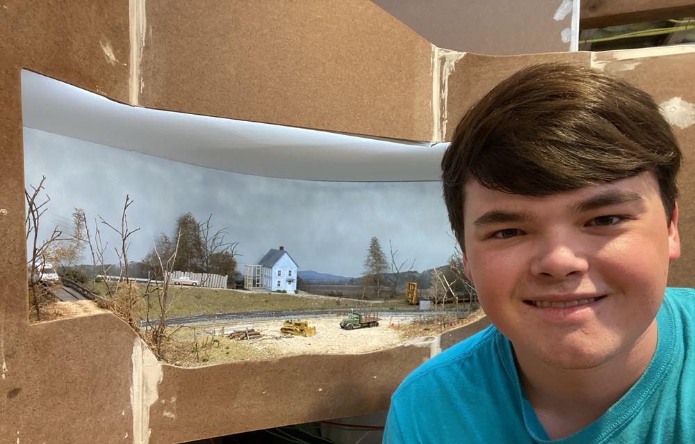 A college-aged boy sits in front of a train layout