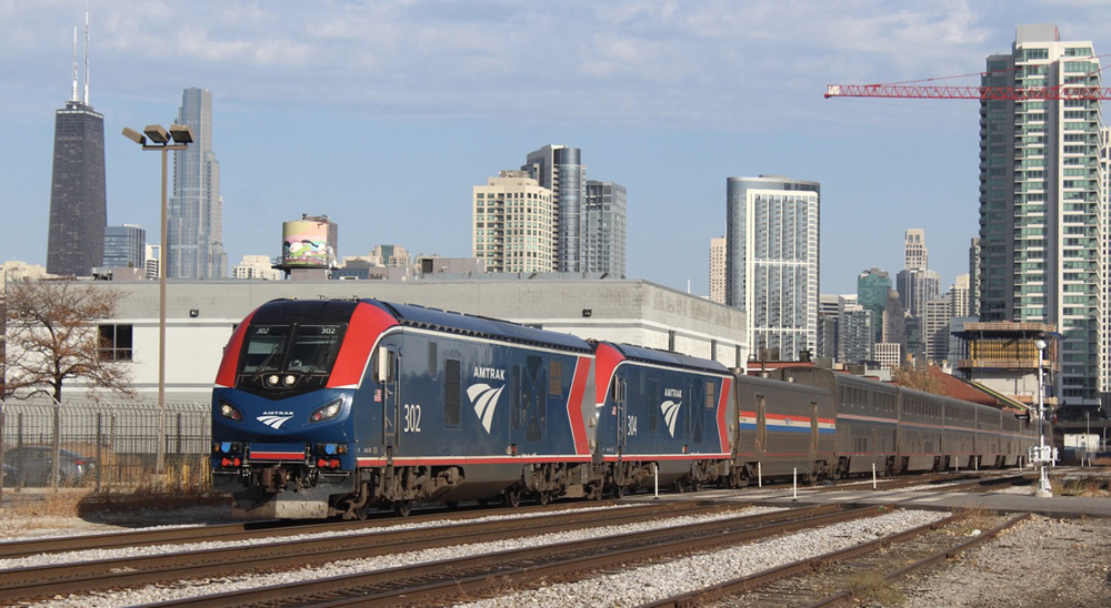 Two blue and red locomotives on passenger train with Chicago skyline in background