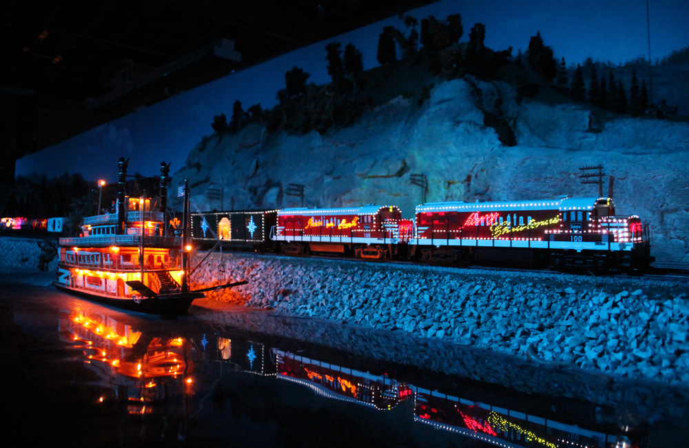 A train decorated with Christmas lights rolls along a riverbank at night, past a brightly lighted riverboat