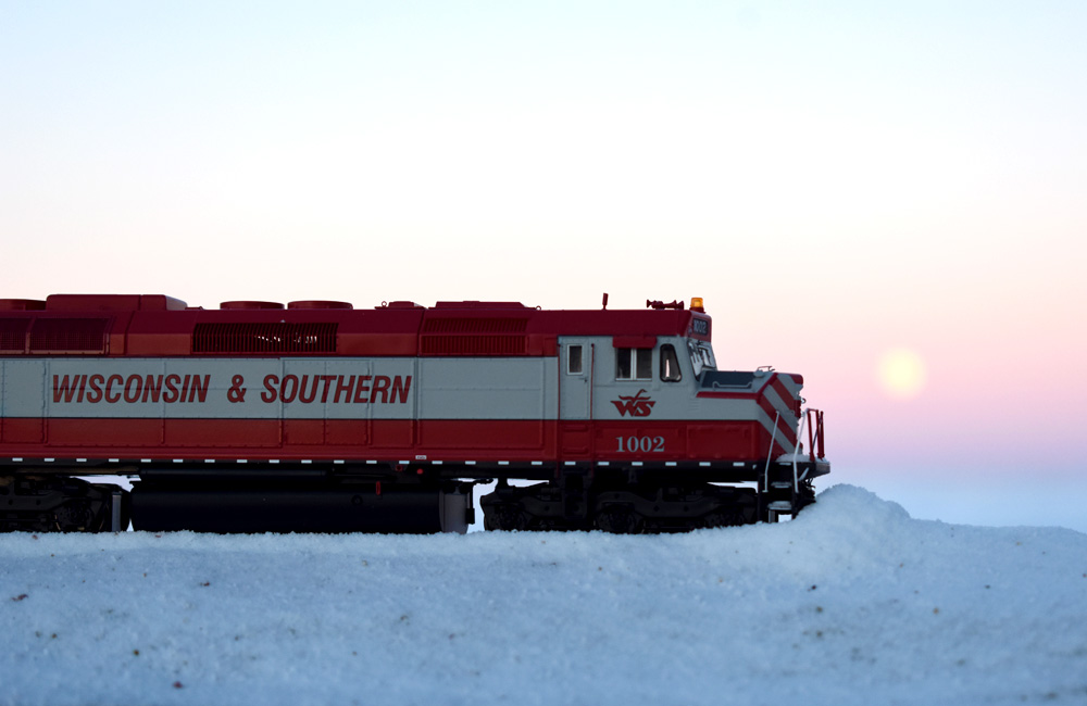 A red-and-white diesel in profile pushes snow off the tracks as the sun rises behind it