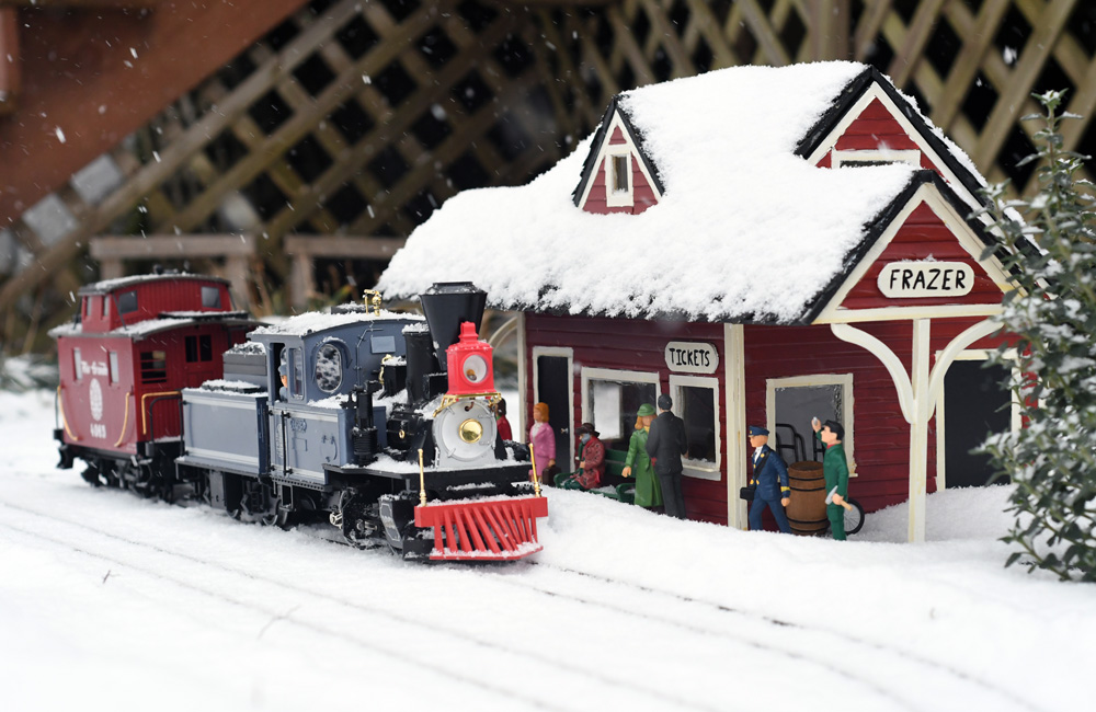 A blue 4-4-0 steam locomotive and its caboose sit at a small wooden depot as snow falls