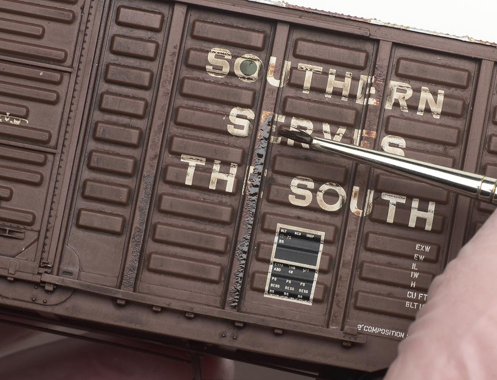 Weathering with artist's oils : A brush is used against the side of a briown model boxcar to weather the doors.