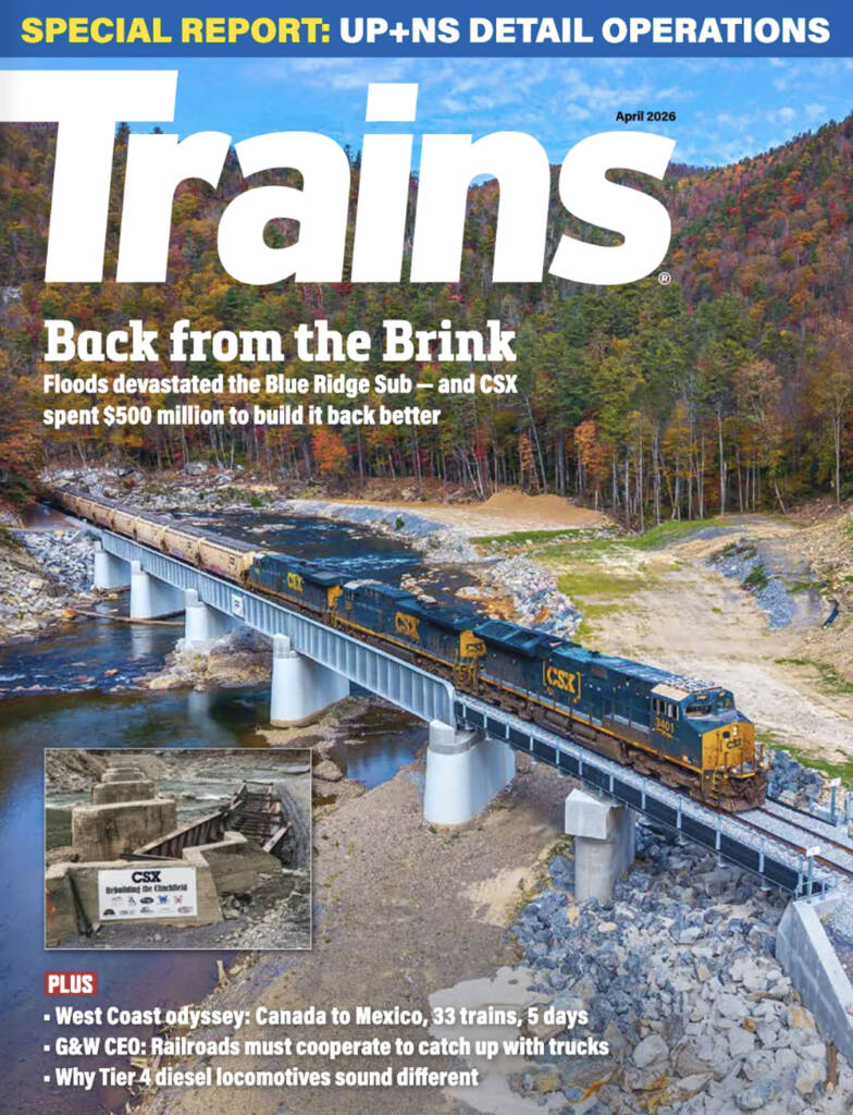 The April 2026 cover of Trains Magazine featuring a freight train crossing a bridge spanning a river in a forest