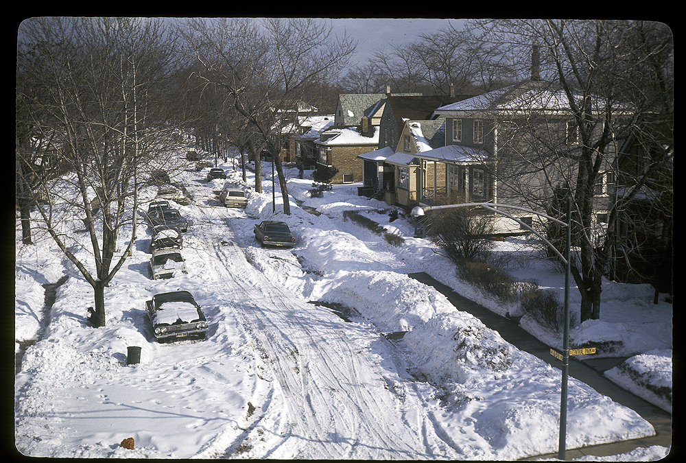 Snow-in streets with cars and partly shoveled sidwalks.