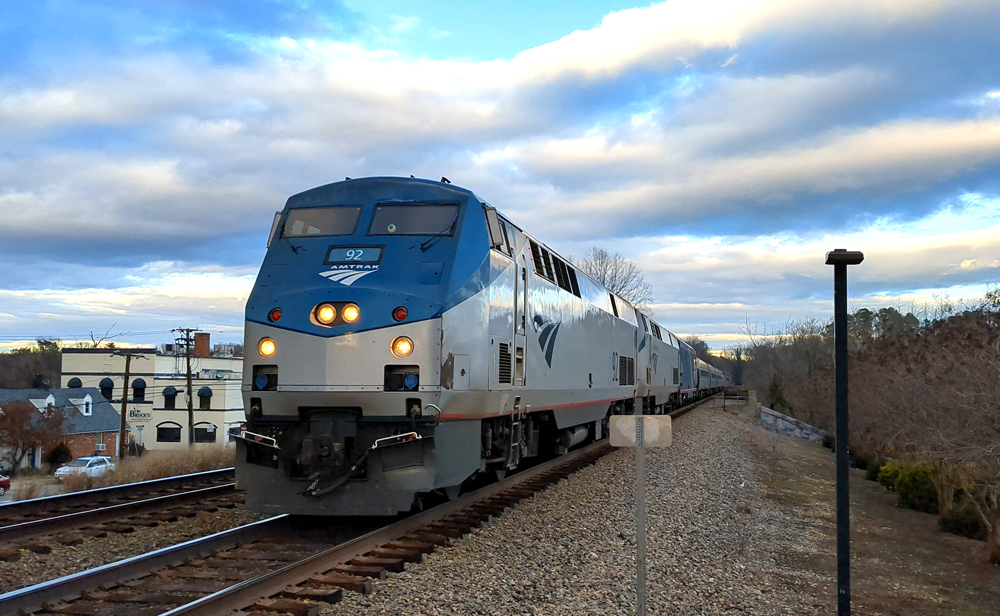Passenger train with three locomotives arrives at station under partly cloudy skies