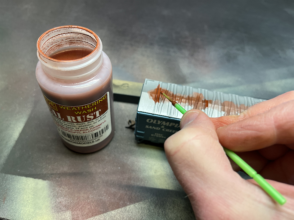 Hand holding green paint brush applying bright orange rust solution to silver model