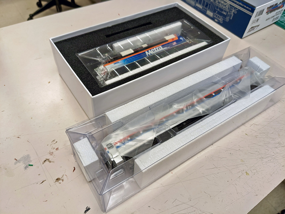 A model of a diesel locomotive sits in a plastic blister surrounded by foam while a passenger car sits in a plastic blister by itself.