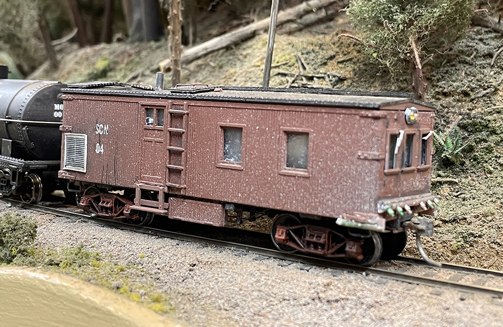 Small model of a brown railroad car with large windows on mountain side railroad track.