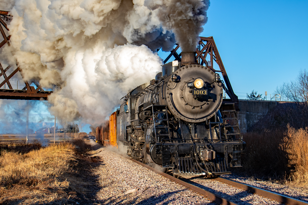 Restored Soo Line Mikado-type steam engine rolls under a rusty railroad overpass