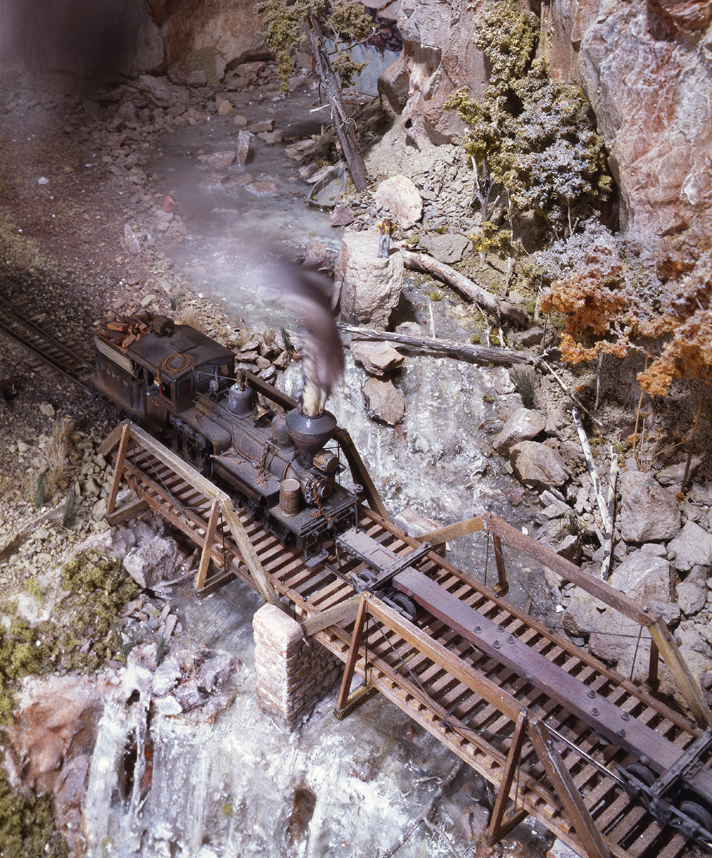 Malcolm Furlow's waterfall modeling technique: Model train on wood bridge over rushing waterfall in rugged mountain scene.