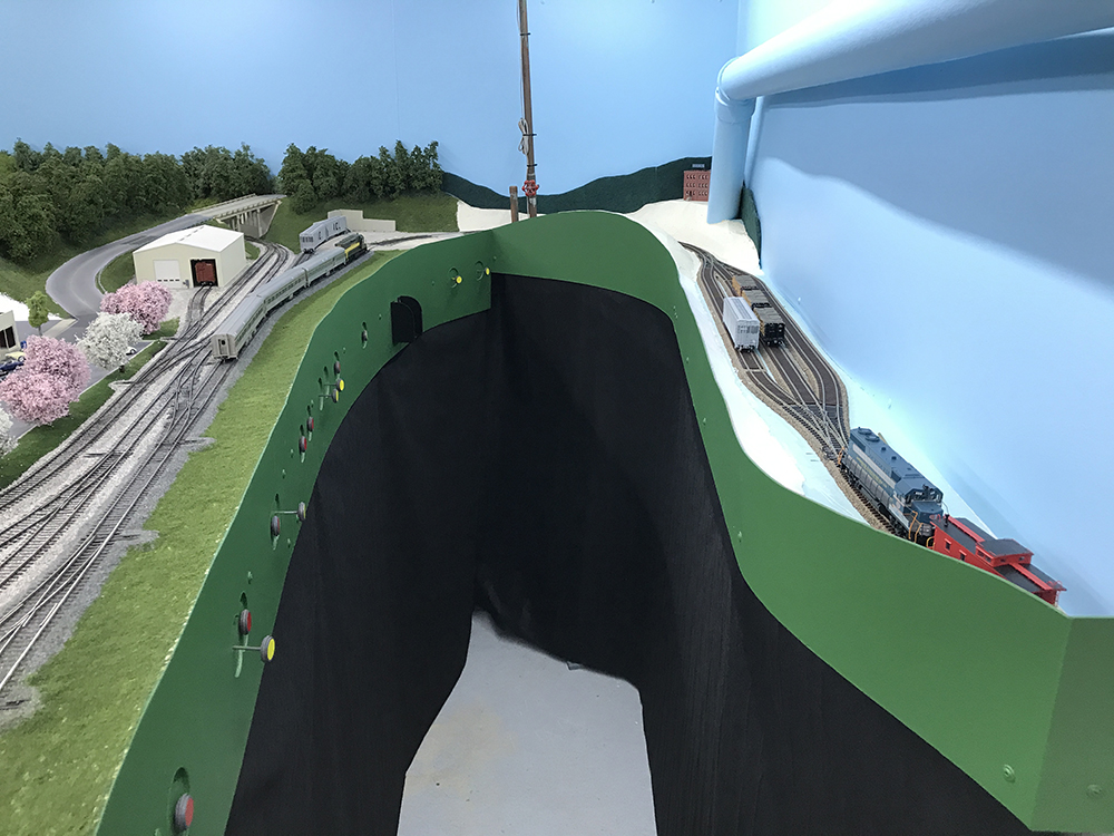 Aisleway for a model train layout with blue sky, green train table, black curtains to hide the support legs, and gray flooring