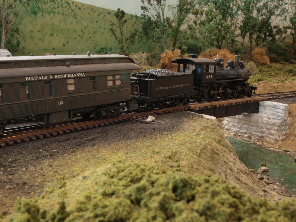 A model steam locomotive pulls a passenger car over a bridge.