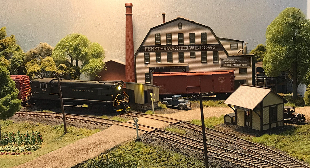 Black model locomotive on model train set with green trees and grass and white buildings