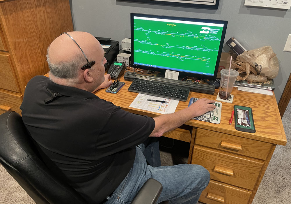 A man in a black shirt sits at a desk and operates a computer that displays a railroad route schematic