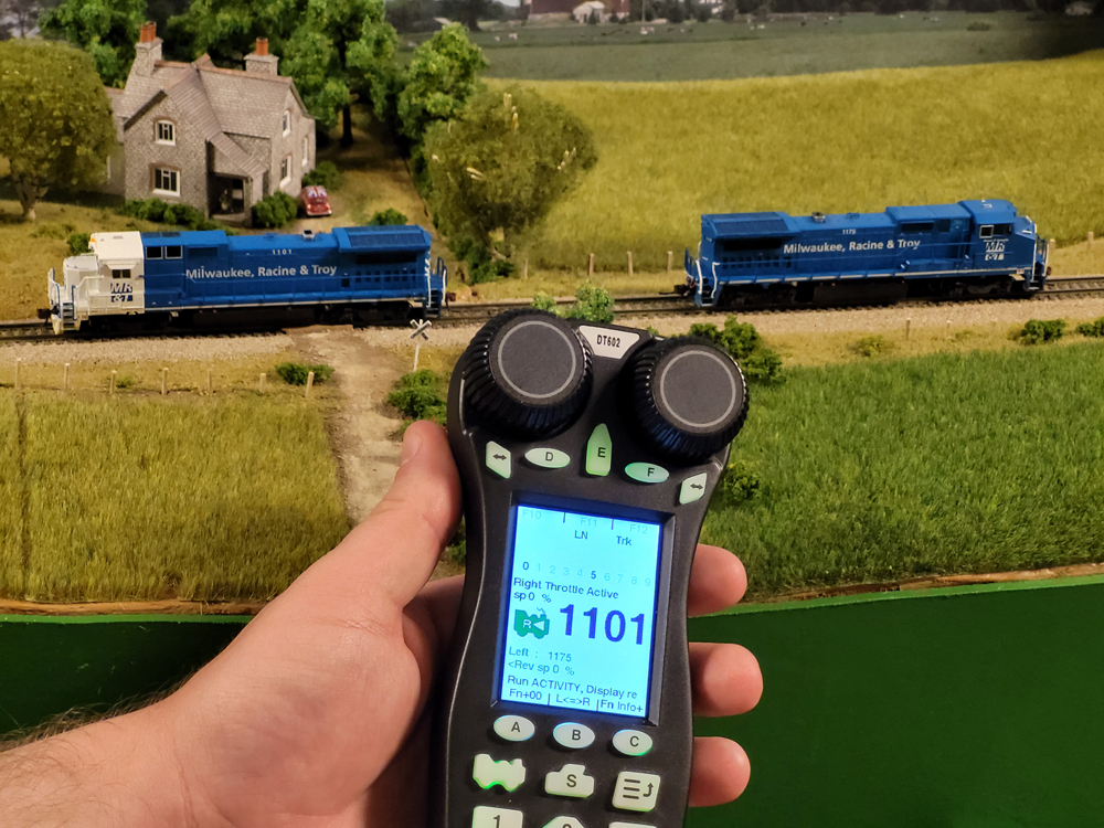 A man holds a model train throttle in front of two blue model locomotives.