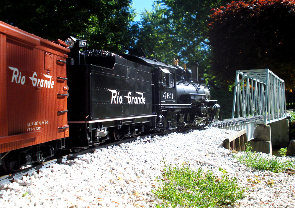 model steam locomotive with two cars head toward bridge on garden railway
