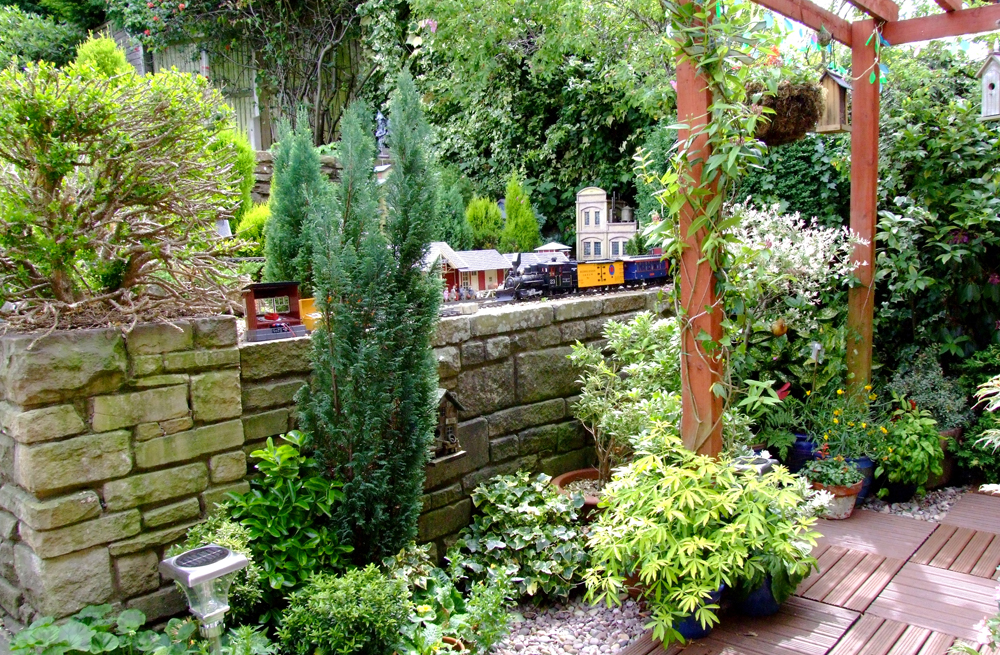 scene on garden railway with greenery, pergola, and a stone retaining wall
