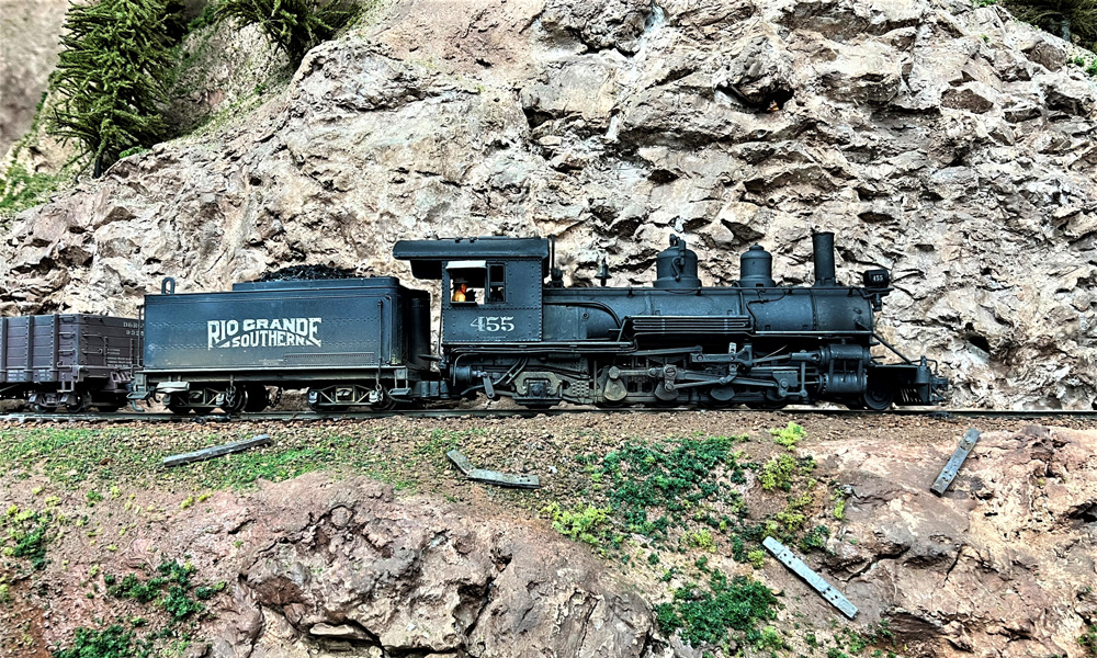 An outside-frame steam locomotive pulls a gondola past a rock wall
