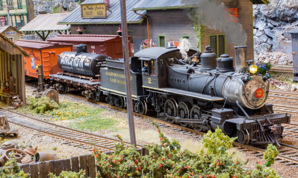 A small steam engine pulls a black tank car and orange refrigerated car past a brick freight house