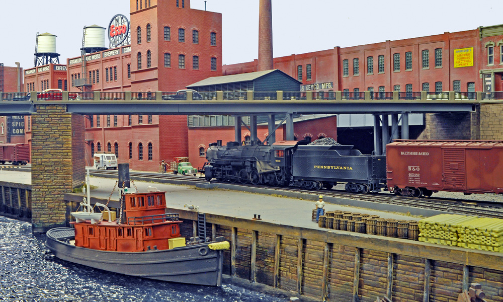 A tugboat bobs alongside a wharf as a steam engine pulls a boxcar through a waterfront industrial scene