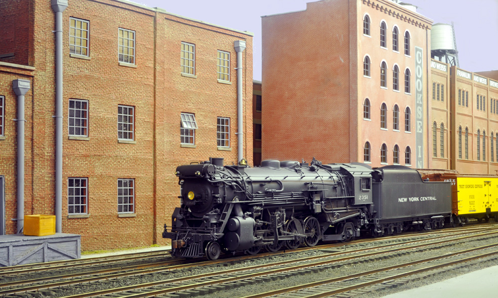 A black steam locomotive pulls a train past a row of brick industrial buildings