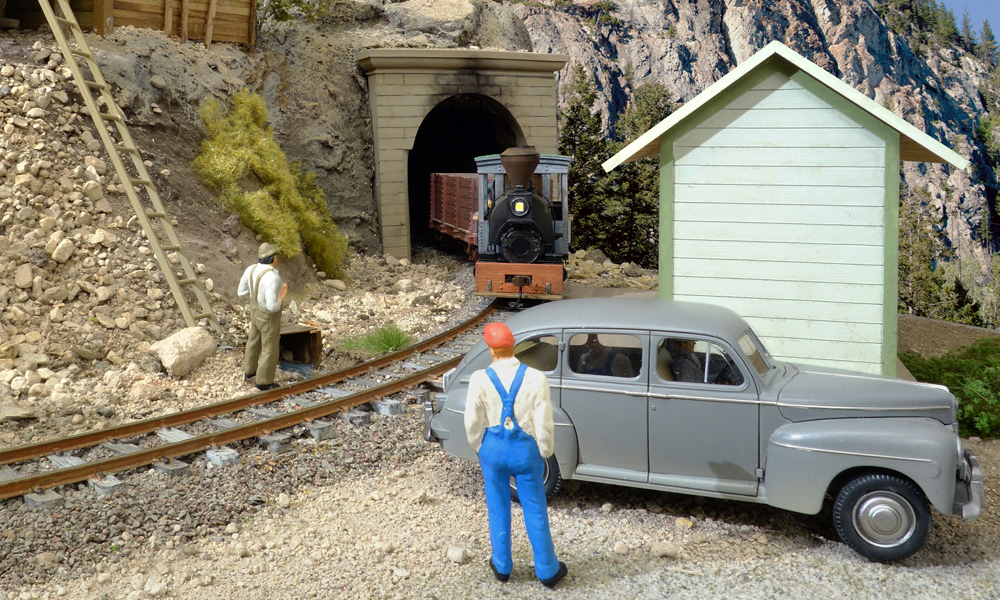 A short steam locomotive pulling gondolas emerges from a tunnel