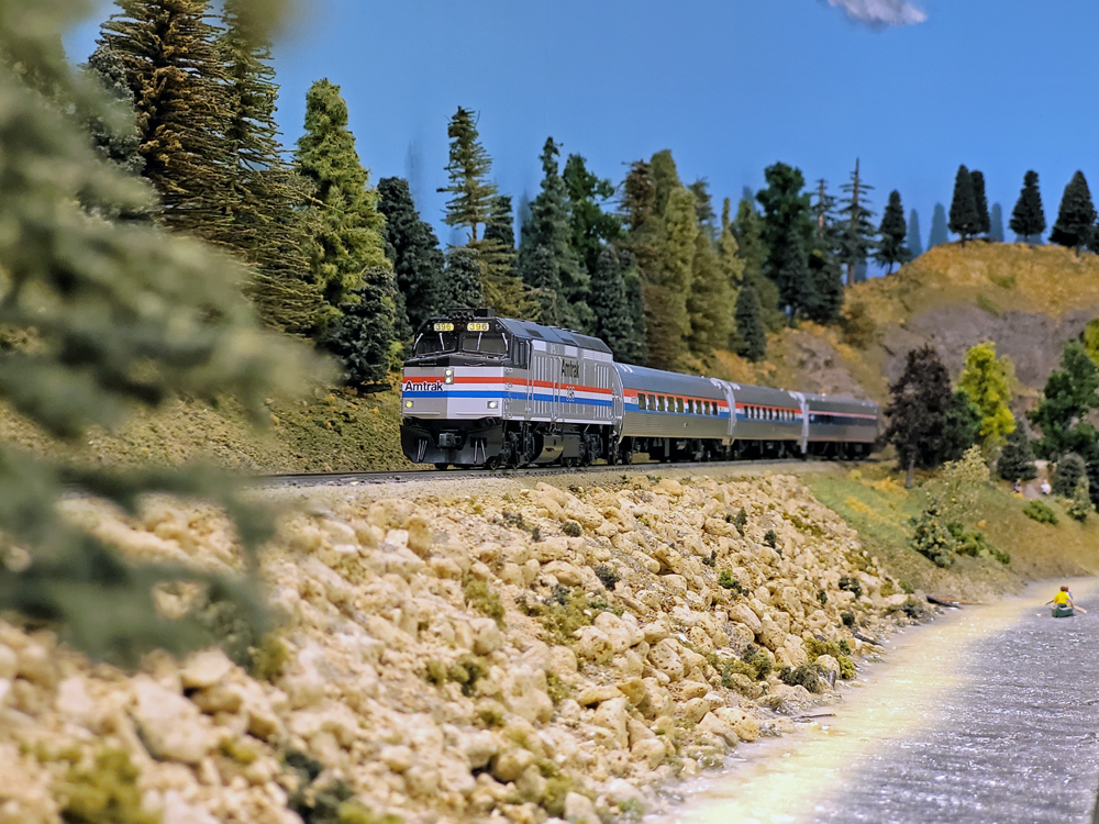 A model diesel locomotive pulls a passenger train along a waterfront.