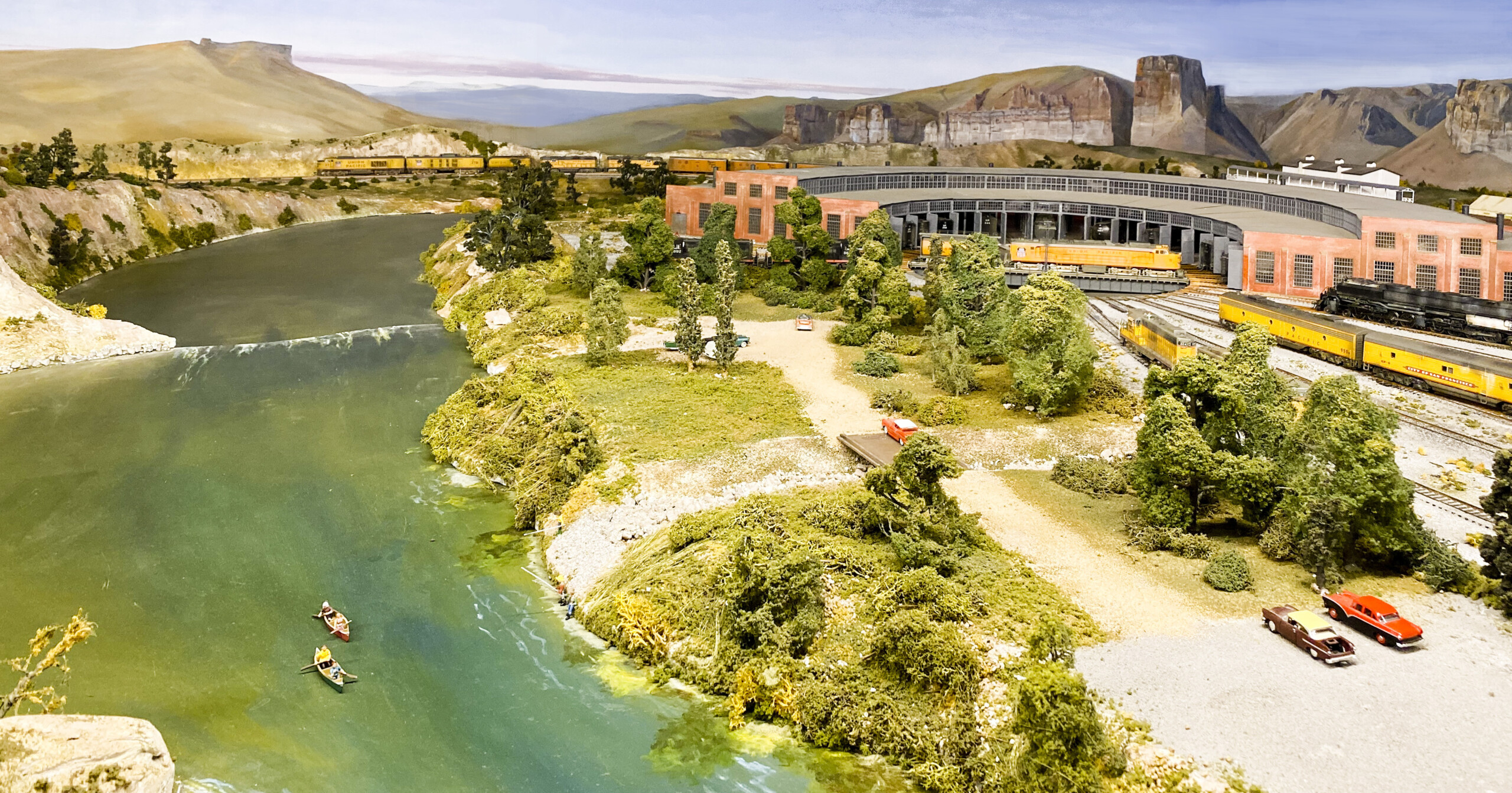 The Green River layout in HO scale: An image of a model railroad layout, with a river on the left of the image, and a locomotive roundhouse on the right, with a desert background