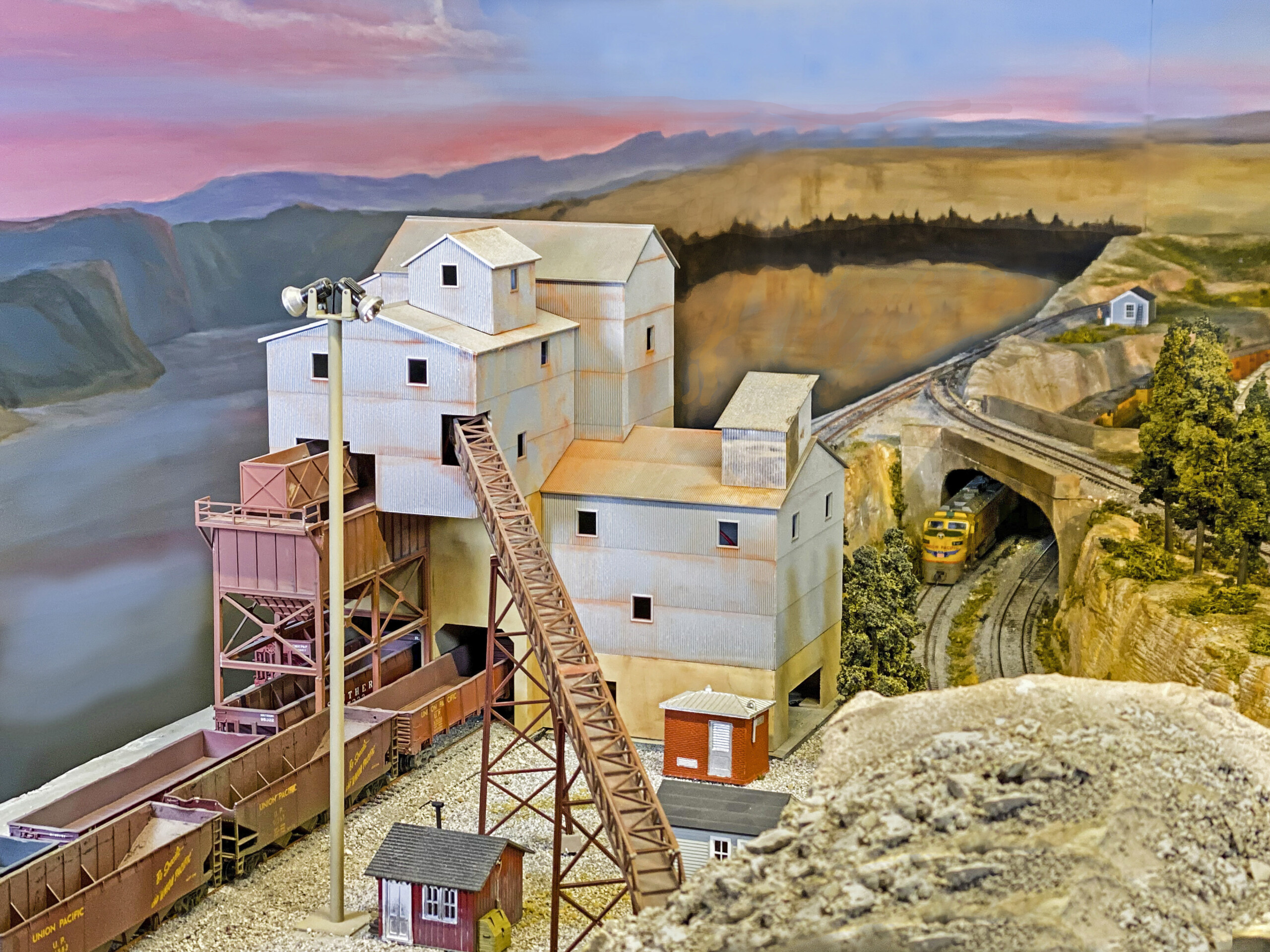 An elevated ballast factory structure is shown with a train passing beneath it in front of a pink sunset
