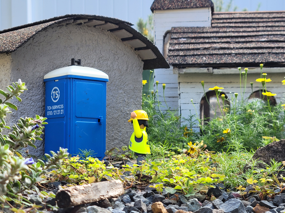 Playmobil figure with arms up next to a model outhouse