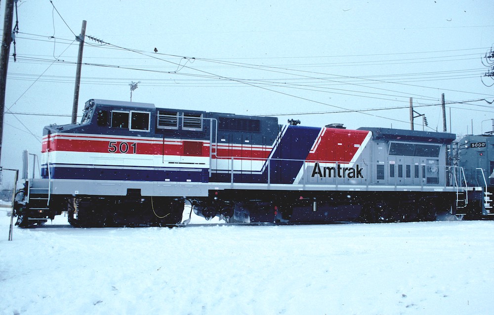 A wintry images of a gray-painted Amtrak locomotive with wide red, white, and blue stripes in a snow storm.