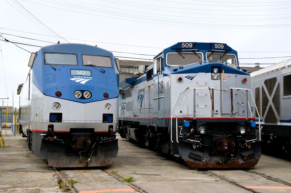 Under overcast skies, two different Amtrak locomotive modes sit side-by-side. Both have gray-painted bodies with blue along the rooflines