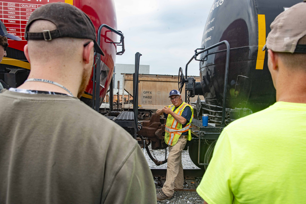 Men watch instructor speaking from between tank cars