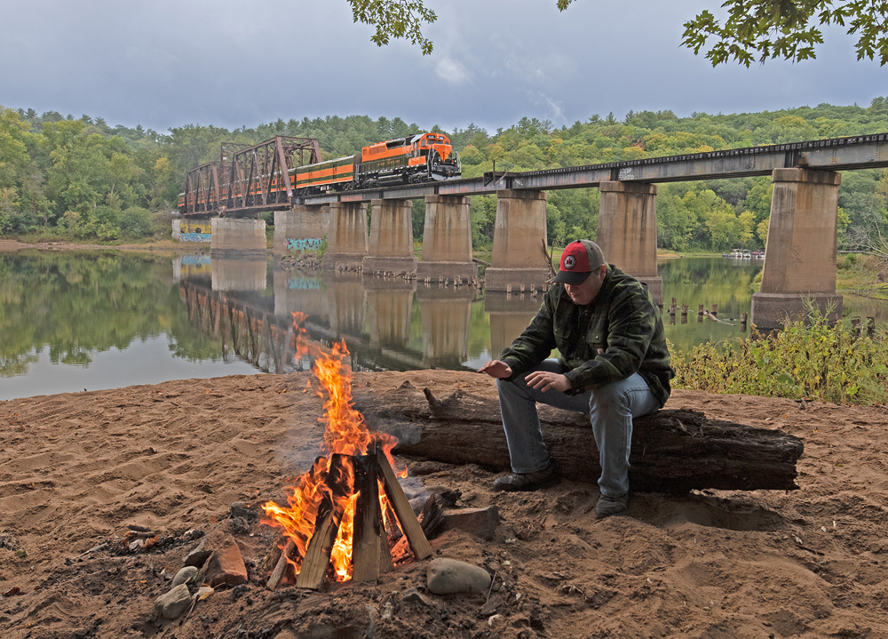 Man sitting by campfire with train in background
