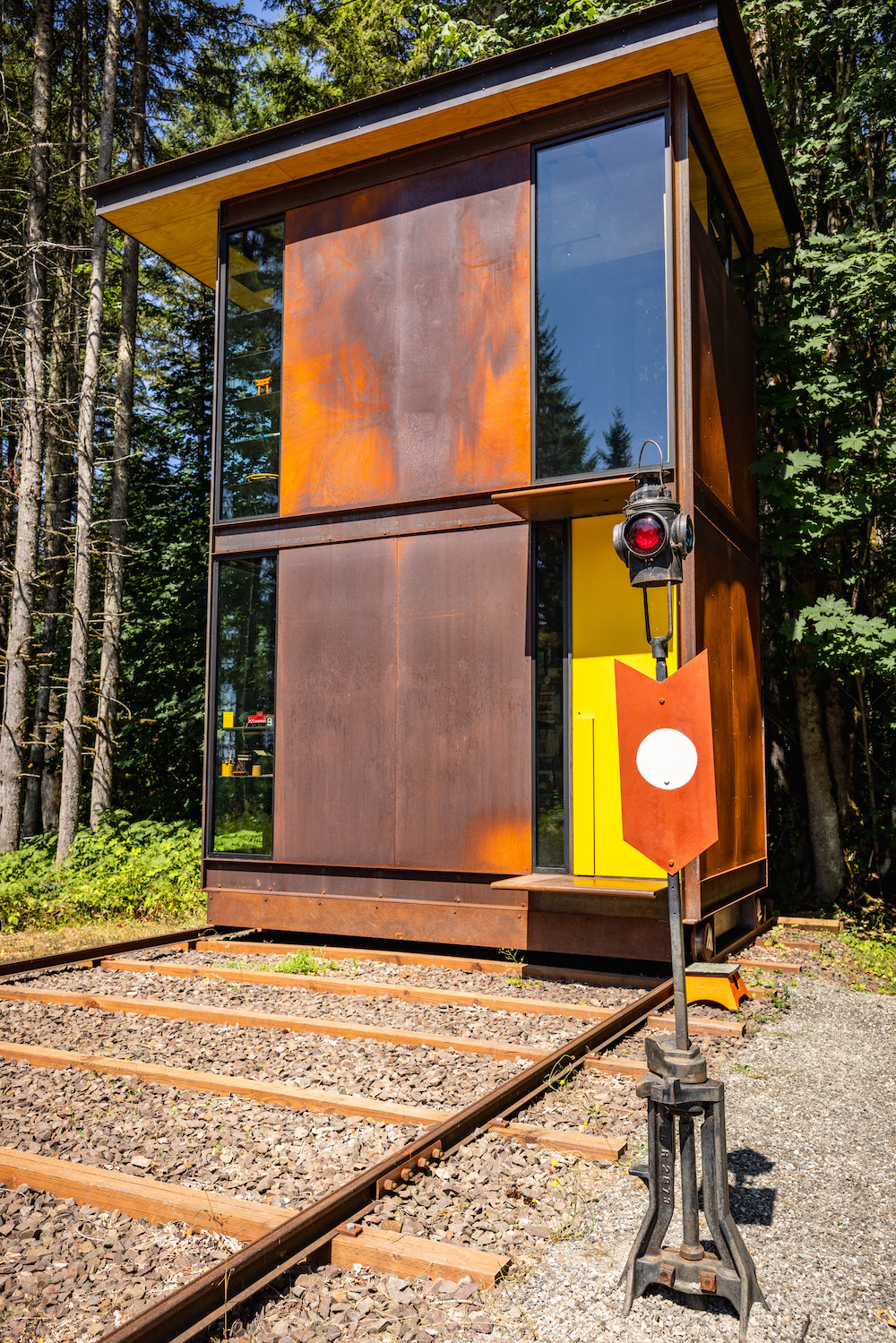 A two-story office structure made of glass and unpainted steel sheets sits on a wide-gauge track