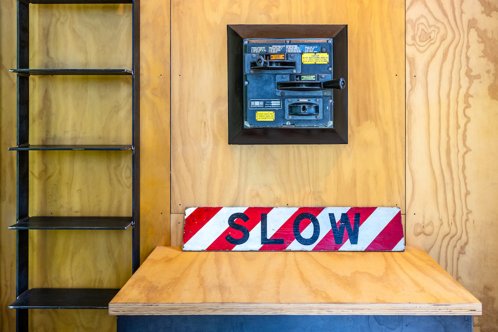 A wall, built of raw plywood sheets with a locomotive control stand and a rad and white striped sign reading slow