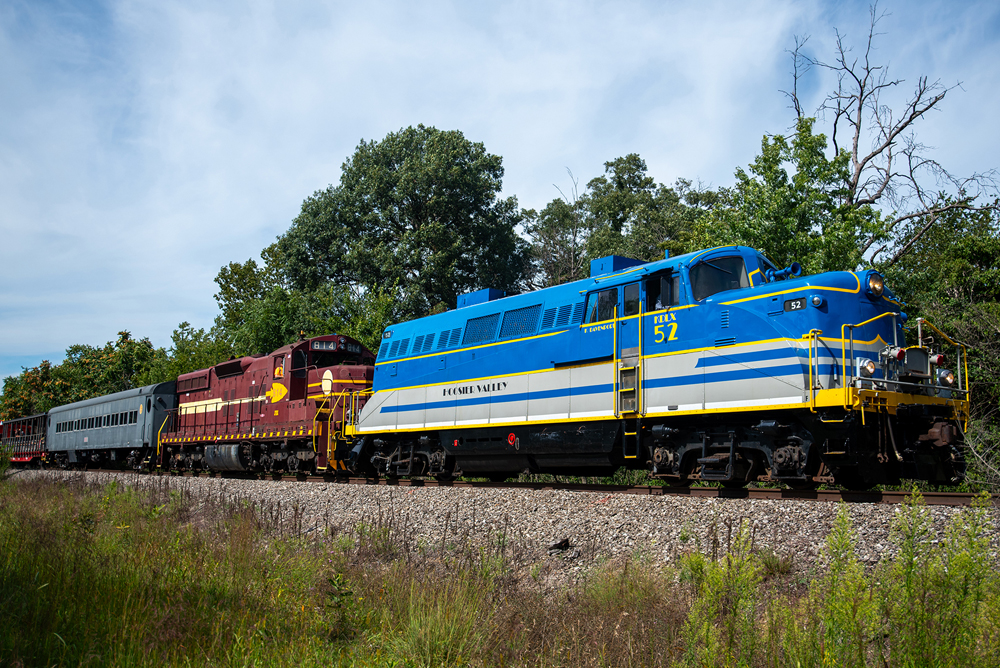 Blue and gray locomotive with yellow trim followed by maroon and yellow locomotive leading passenger train