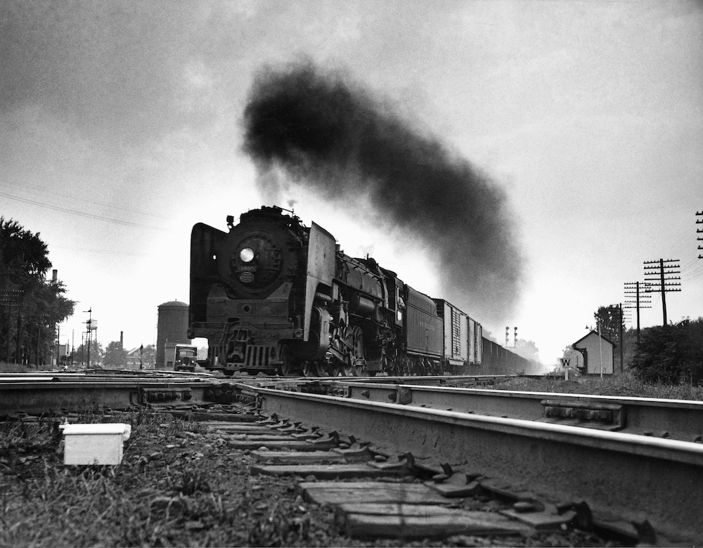 A black and white photograph from 1956 shows a large, black steam engine crossing another set of railroad tracks set at an angle while black coal smoke flls the sky