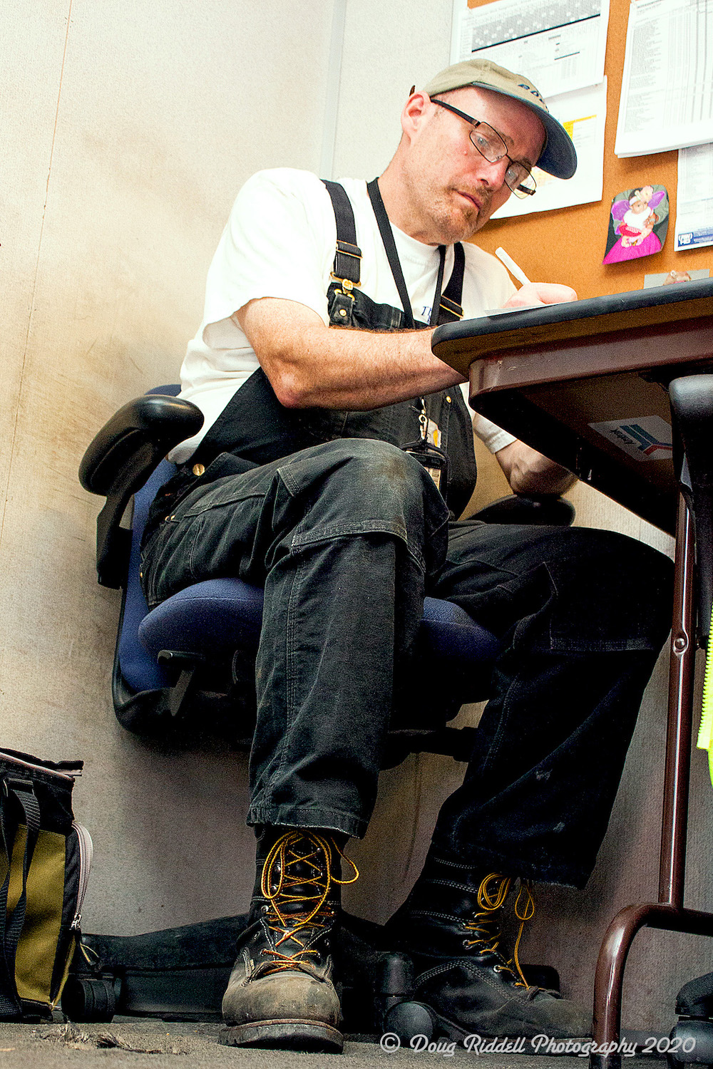 Clad in work boots and a overalls, a railroad worker works on paperwork at a table.