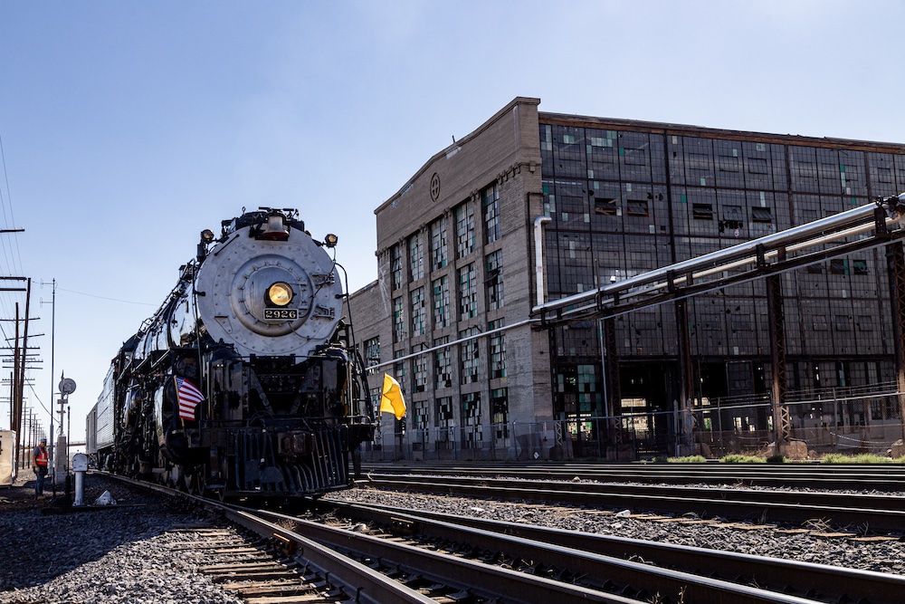 Steam locomotive operates on the main line with a massive facility building in the background.